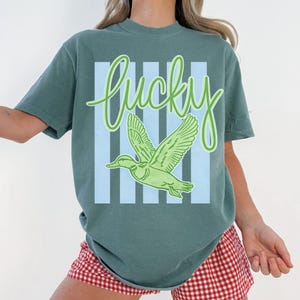 May include: A teal-colored t-shirt with the word "Lucky" in a green script font. Below the word is a green duck graphic with outstretched wings, set against a backdrop of light blue vertical stripes. The shirt is a casual style.