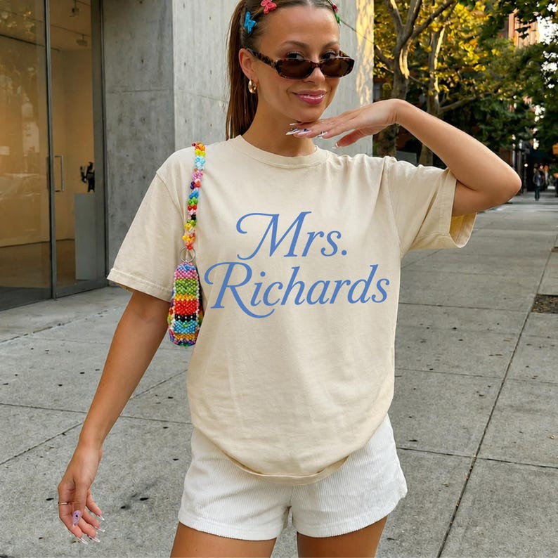 Może przedstawiać: Beżowy t-shirt z kr&oacute;tkim rękawem z niebieskim napisem Mrs. Richards. Koszulka jest zestawiona z białymi szortami i kolorowym paskiem do torebki z koralik&oacute;w. Odzież posiada okrągły dekolt oraz luźny, swobodny kr&oacute;j, kt&oacute;ry zapewnia komfort noszenia.