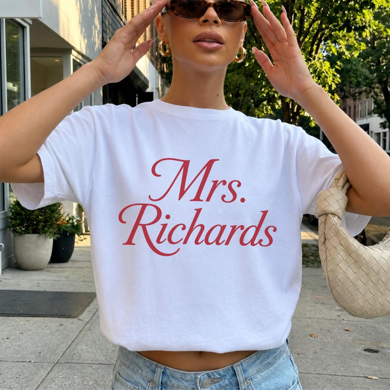 Może przedstawiać: Biały t-shirt z kr&oacute;tkim rękawem z napisem Mrs. Richards nadrukowanym czerwoną kursywą na klatce piersiowej. Koszulka ma okrągły dekolt i standardowy kr&oacute;j, zestawiona z jasnoniebieskimi dżinsami oraz plecioną beżową torebką na ramię.