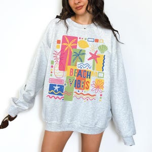 Pu&ograve; includere: Felpa grigio chiaro con un design grafico colorato. Il design include un ananas, palme, stelle marine e la scritta "BEACH VIBES". La felpa &egrave; uno stile casual e confortevole.