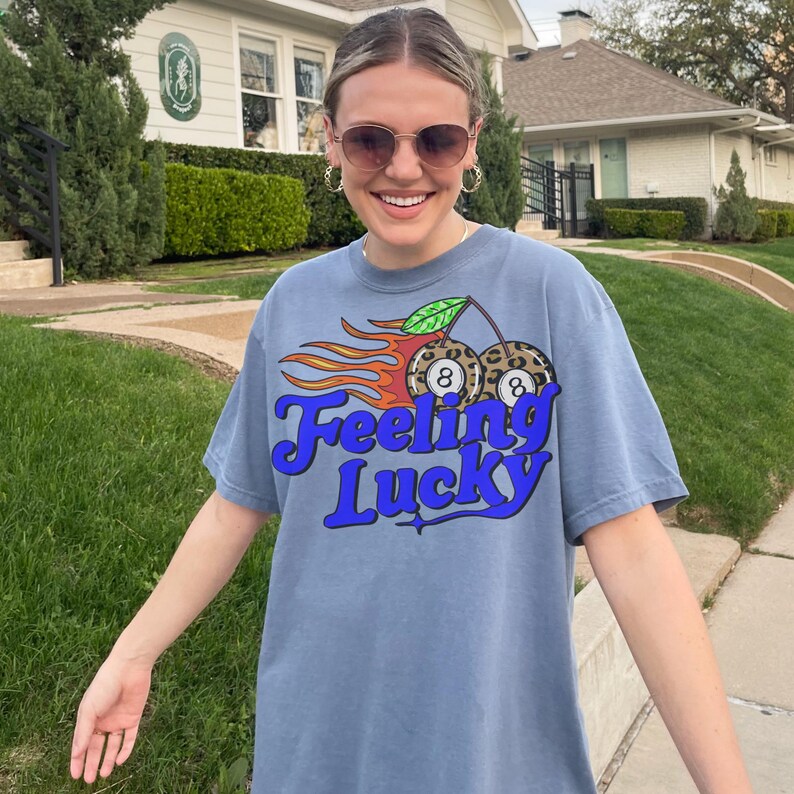 K&ouml;nnte beinhalten: Blaues T-Shirt mit dem Text "Feeling Lucky" in blauer Schrift. Die Grafik zeigt zwei Kirschen mit 8-Ball-Designs und Flammen. Das Shirt ist ein l&auml;ssiger, bequemer Stil.
