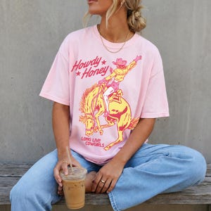 May include: A pale pink t-shirt featuring a cowgirl on a bucking bronco in yellow and red. The text reads "Howdy Honey" and "Long Live Cowgirls". The shirt is worn with blue jeans.