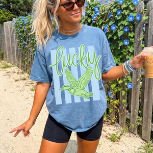 May include: A blue t-shirt with the word "Lucky" in green script and a green duck graphic. The shirt has a light blue striped background. The person is wearing black shorts and sunglasses, and holding a beverage.