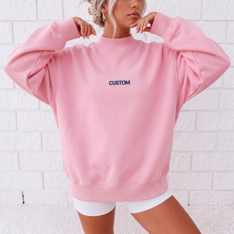 May include: A light pink long-sleeve sweatshirt featuring a mock neck collar and ribbed cuffs. The word CUSTOM is embroidered in dark blue thread across the center of the chest. The garment has a relaxed fit with dropped shoulders and a hem.