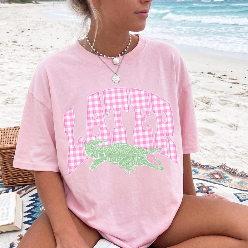 May include: A light pink t-shirt with the word "LATER" in a pink and white gingham pattern. Below the text is a green alligator graphic. The shirt is a casual style, perfect for a day at the beach.