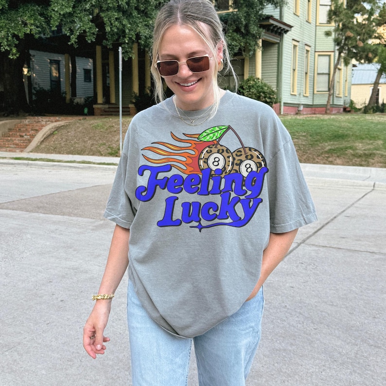 K&ouml;nnte beinhalten: Graues T-Shirt mit dem Text "Feeling Lucky" in blauer Schrift. Die Grafik zeigt zwei Billardkugeln mit der Nummer 8, Flammen und ein Kirschdesign. Das Shirt hat eine lockere, &uuml;bergro&szlig;e Passform.