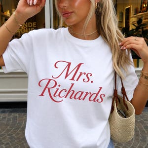 Może przedstawiać: Biały t-shirt z kr&oacute;tkim rękawem i okrągłym dekoltem, z napisem Mrs. Richards nadrukowanym czerwoną kursywą na środku klatki piersiowej. Odzież wykonana jest z gładkiej białej tkaniny i ma standardowy kr&oacute;j z okrągłym wycięciem pod szyją.