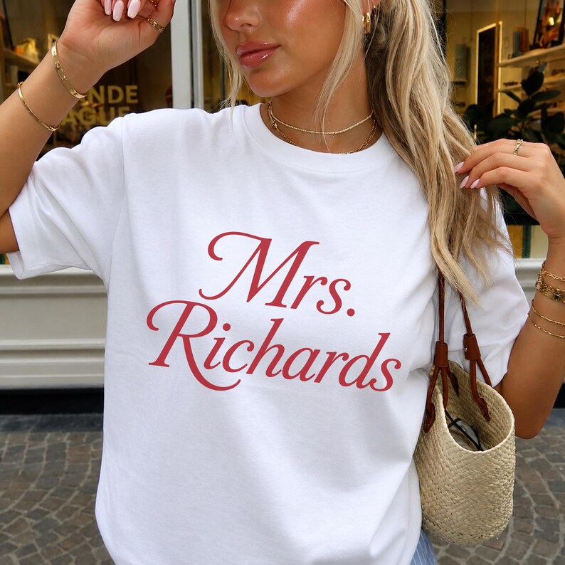 Może przedstawiać: Biały t-shirt z kr&oacute;tkim rękawem i okrągłym dekoltem, z napisem Mrs. Richards nadrukowanym czerwoną kursywą na środku klatki piersiowej. Odzież wykonana jest z gładkiej białej tkaniny i ma standardowy kr&oacute;j z okrągłym wycięciem pod szyją.
