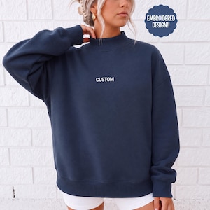 May include: A dark navy blue long-sleeve sweatshirt with a mock neck collar. The word CUSTOM is embroidered in white thread across the center of the chest. A dark blue scalloped badge in the upper right corner contains the text EMBROIDERED DESIGN!!