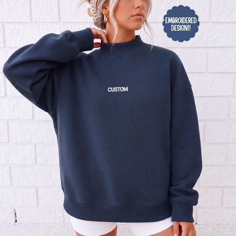 May include: A dark navy blue long-sleeve sweatshirt with a mock neck collar. The word CUSTOM is embroidered in white thread across the center of the chest. A dark blue scalloped badge in the upper right corner contains the text EMBROIDERED DESIGN!!