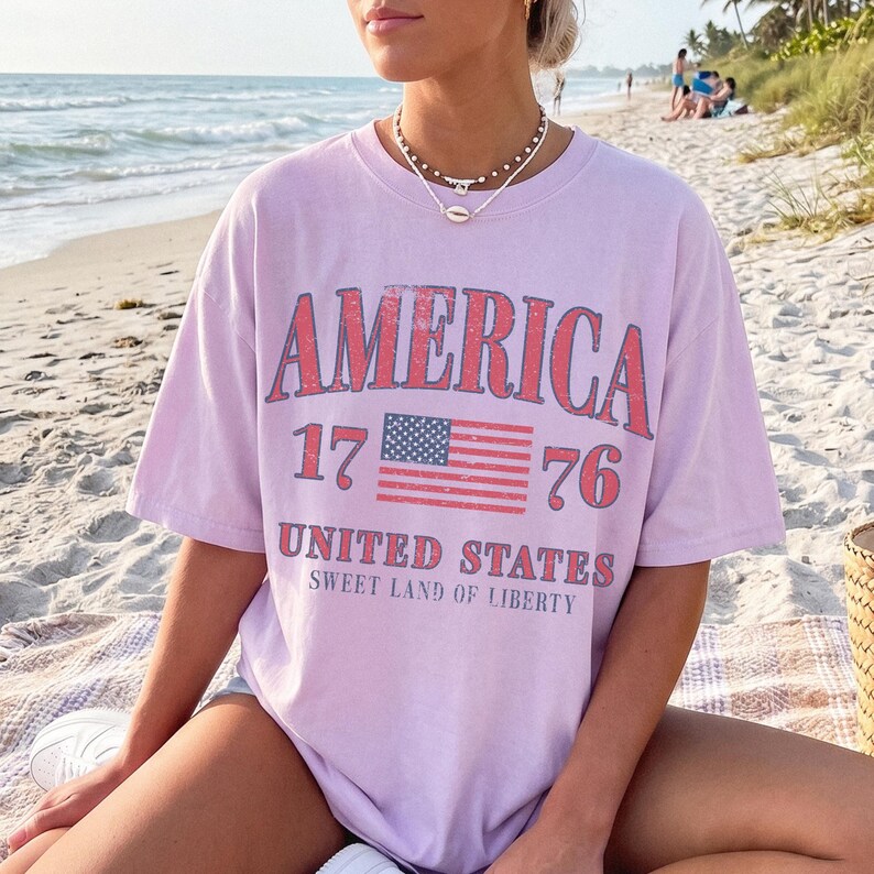 May include: A lilac t-shirt featuring the word "AMERICA" in red, the numbers "17" and "76", and a red and blue American flag graphic. The text "UNITED STATES" and "SWEET LAND OF LIBERTY" are below the flag.