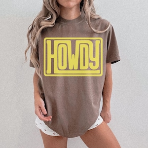 May include: A brown t-shirt featuring the word "HOWDY" in a yellow, retro font, within a yellow rectangular frame. The shirt has a relaxed fit and a neutral colour. The text is clearly visible.