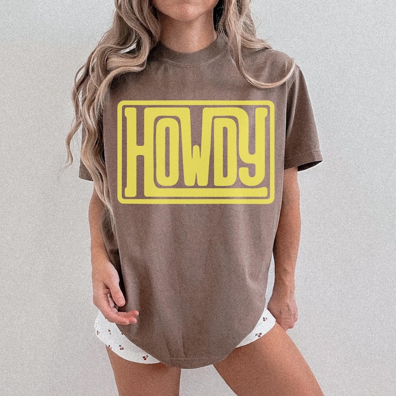 May include: A brown t-shirt featuring the word "HOWDY" in a yellow, retro font, within a yellow rectangular frame. The shirt has a relaxed fit and a neutral colour. The text is clearly visible.