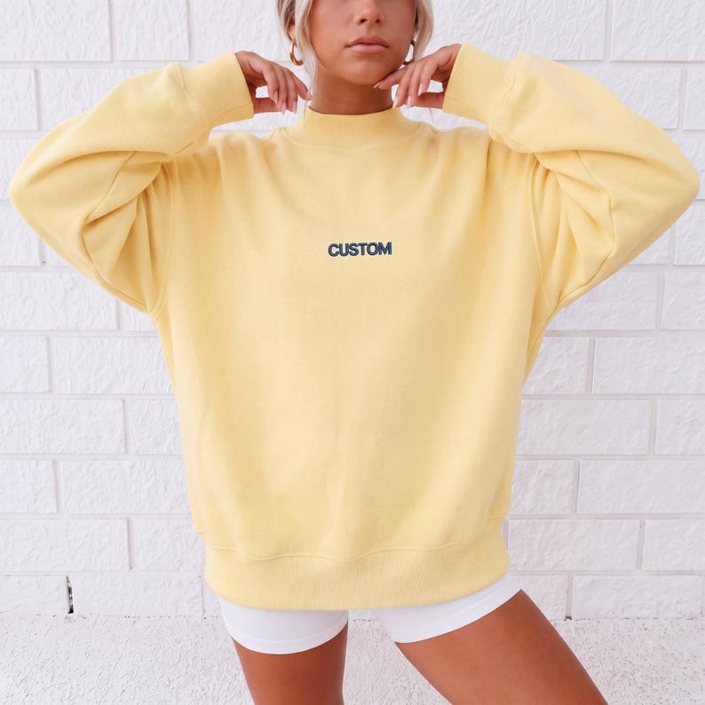 May include: A pale yellow long-sleeve sweatshirt featuring a mock neck collar and ribbed cuffs. The word CUSTOM is embroidered in dark blue capital letters across the center of the chest. The garment has a relaxed fit with dropped shoulders.