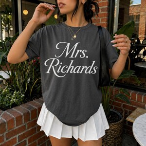 Może przedstawiać: Ciemnoszary t-shirt z kr&oacute;tkim rękawem z białym napisem Mrs. Richards wydrukowanym kursywą na klatce piersiowej, w połączeniu z białą plisowaną sp&oacute;dnicą i czarną torbą na ramię z cienkim paskiem.