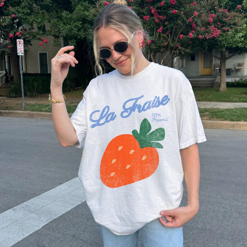 May include: Cream-coloured t-shirt featuring a large orange strawberry illustration and the blue text "La Fraise". The shirt also has the text "100% Organic". The t-shirt is short-sleeved.