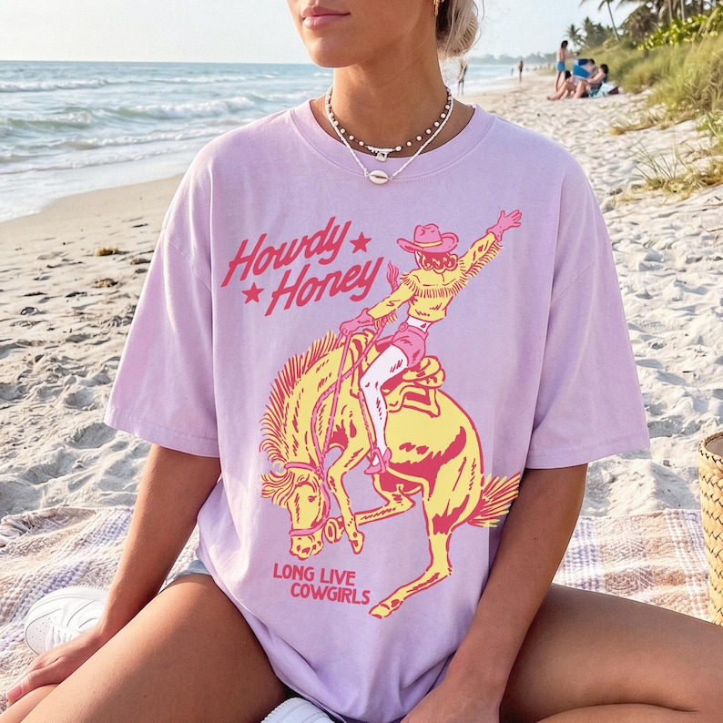May include: A lilac t-shirt featuring the text "Howdy Honey" and a graphic of a cowgirl riding a bucking bronco. The words "Long Live Cowgirls" are printed below the graphic. The shirt is worn on a beach.