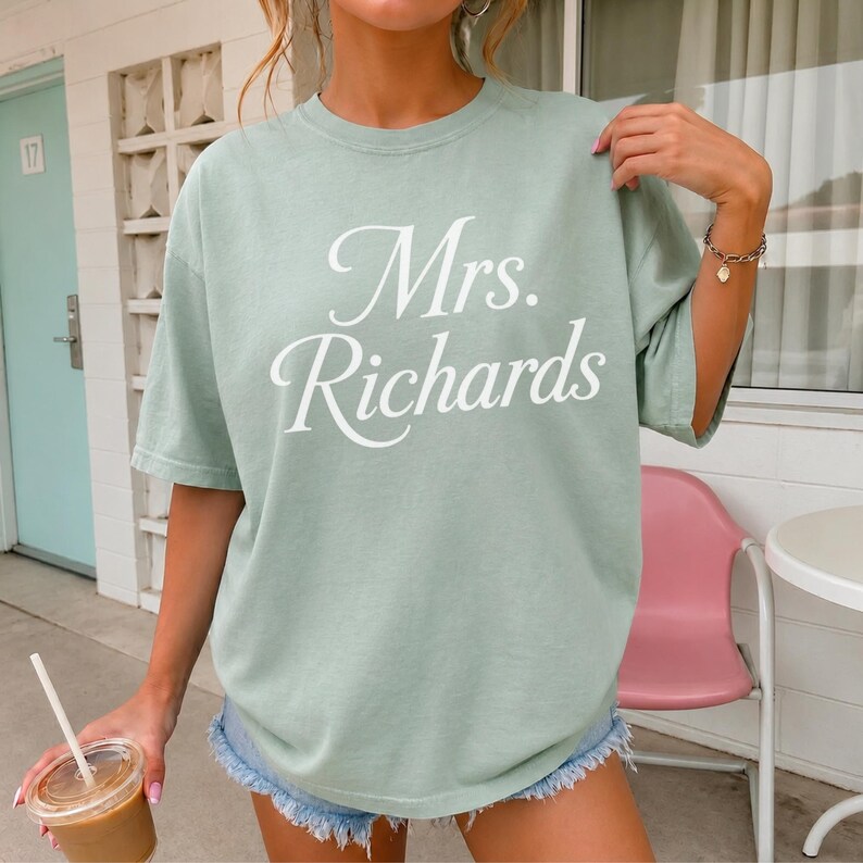 Może przedstawiać: Szałwiowo-zielony t-shirt z kr&oacute;tkim rękawem z białym napisem Mrs. Richards wydrukowanym kursywą na klatce piersiowej. Koszulka ma okrągły dekolt i luźny kr&oacute;j, zestawiona z jasnoniebieskimi szortami dżinsowymi z postrzępionym wykończeniem.