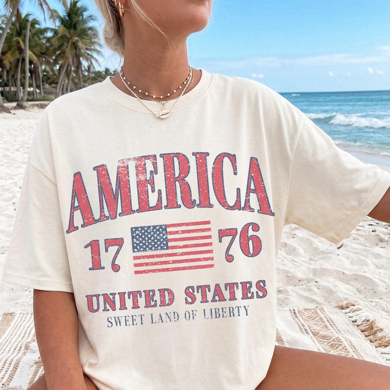 May include: Cream-coloured t-shirt featuring the word "AMERICA" in distressed red lettering. Below, the number "1776" and a flag graphic are displayed. The text "UNITED STATES" and "SWEET LAND OF LIBERTY" are also visible.