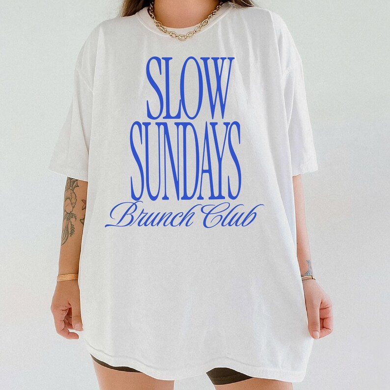 Peut inclure: T-shirt blanc avec l'inscription bleue "SLOW SUNDAYS Brunch Club". Le texte est centr&eacute; sur le devant. Le t-shirt a des manches courtes et une coupe ample, id&eacute;al pour le brunch.