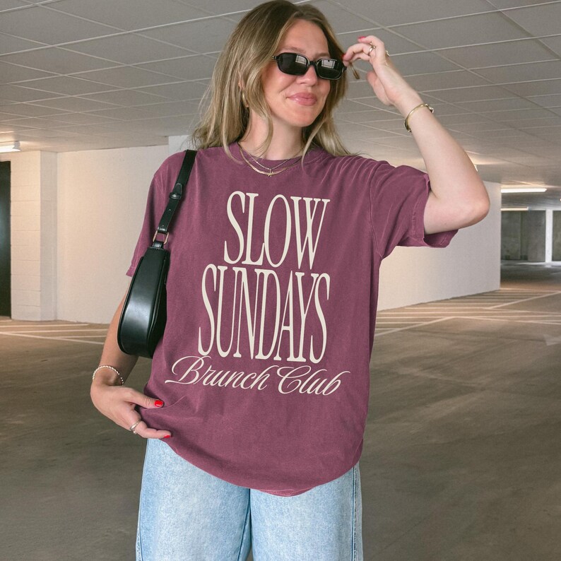Peut inclure: T-shirt bordeaux avec l'inscription "SLOW SUNDAYS Brunch Club" en blanc. Le t-shirt est associ&eacute; &agrave; un jean bleu clair et un sac &agrave; bandouli&egrave;re noir. La personne porte des lunettes de soleil noires.