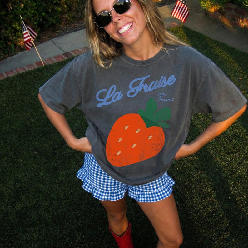 May include: Dark grey t-shirt with a large orange strawberry graphic and the text "La Fraise 100% Organic". The person is wearing blue and white checked shorts and red boots. Sunglasses are worn.