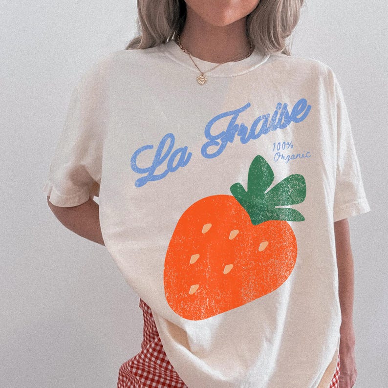 May include: Cream-coloured t-shirt with a large orange strawberry graphic and green leaves. The text "La Fraise" in blue script and "100% Organic" is printed above the strawberry. The shirt has short sleeves.