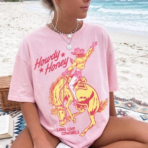 May include: Pale pink t-shirt featuring a cowgirl on a bucking bronco in yellow and red. The text "Howdy Honey" and "Long Live Cowgirls" are printed on the shirt. The t-shirt has a relaxed, oversized fit.