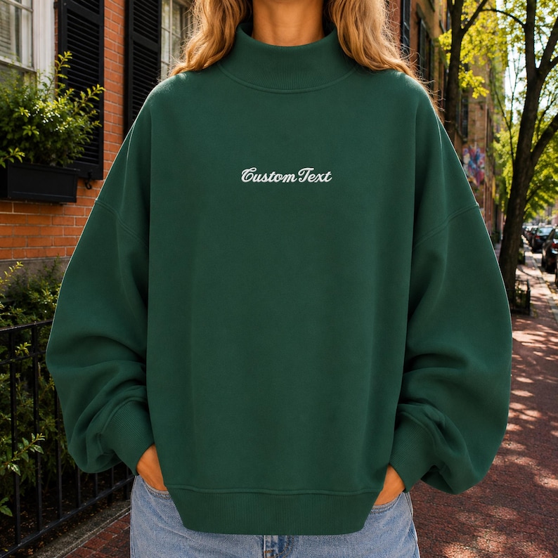 May include: A forest green mock neck sweatshirt featuring long sleeves and a ribbed hem. The center of the chest displays the text CustomText embroidered in a white cursive font. The garment has a relaxed fit with dropped shoulders and ribbed cuffs.
