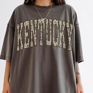 May include: Dark grey t-shirt with the word "KENTUCKY" in large, block letters across the front. The letters have a camouflage pattern in shades of brown and green. The shirt has short sleeves and a relaxed fit.