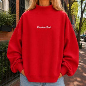 May include: A bright red long-sleeve sweatshirt with a mock neck collar. The center of the chest features the white embroidered text CustomText in a cursive script font. The garment has a relaxed fit with dropped shoulders and ribbed cuffs.