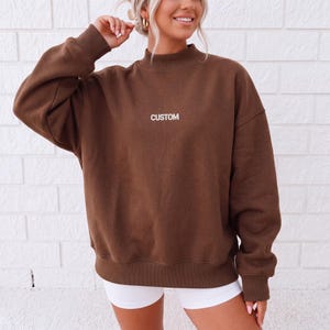May include: A dark brown long-sleeve sweatshirt featuring a mock neck collar and ribbed cuffs. The word CUSTOM is printed in white capital letters on the center of the chest. The garment has a relaxed fit with dropped shoulders and a straight hem.