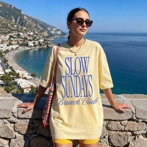 Peut inclure: Un t-shirt jaune clair avec l'inscription bleue "SLOW SUNDAYS Brunch Club". La personne porte des lunettes de soleil et un collier. L'arri&egrave;re-plan montre une vue c&ocirc;ti&egrave;re avec des montagnes et la mer.
