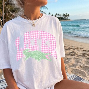 May include: White t-shirt featuring the word "LATER" in pink gingham print above a green alligator graphic. The shirt is worn on a beach with a blue ocean and palm trees in the background.
