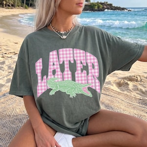 May include: An olive green t-shirt with the word "LATER" in pink gingham print and a green alligator graphic. The shirt is a casual, oversized style, suitable for everyday wear. The image is taken on a beach with the ocean in the background.