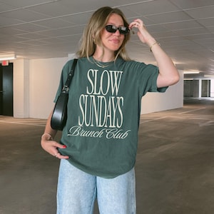 Peut inclure: T-shirt vert fonc&eacute; avec l'inscription blanche "SLOW SUNDAYS Brunch Club". Le t-shirt est associ&eacute; &agrave; un jean bleu clair et un sac &agrave; bandouli&egrave;re noir. La personne porte des lunettes de soleil noires.