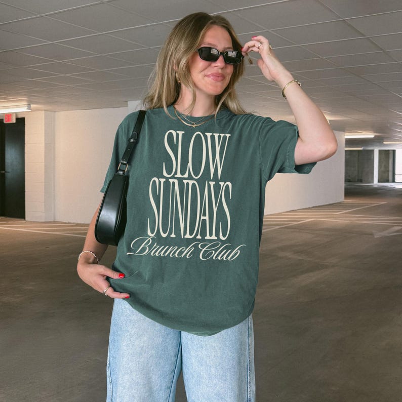 Peut inclure: T-shirt vert fonc&eacute; avec l'inscription blanche "SLOW SUNDAYS Brunch Club". Le t-shirt est associ&eacute; &agrave; un jean bleu clair et un sac &agrave; bandouli&egrave;re noir. La personne porte des lunettes de soleil noires.