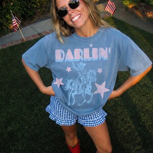 Puede incluir: Camiseta azul con la palabra "DARLIN'" en rosa y azul, con un vaquero a caballo y estrellas rosas. La persona lleva un pantal&oacute;n corto a cuadros azul y blanco y botas rojas.