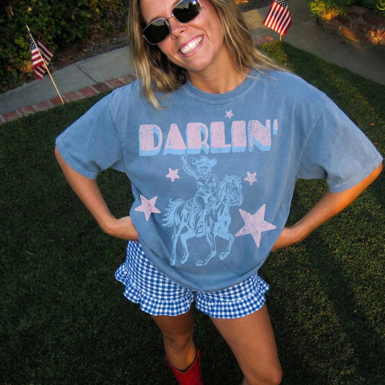 Puede incluir: Camiseta azul con la palabra "DARLIN'" en rosa y azul, con un vaquero a caballo y estrellas rosas. La persona lleva un pantal&oacute;n corto a cuadros azul y blanco y botas rojas.