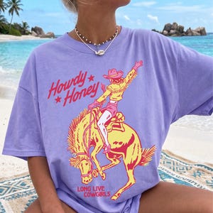 May include: A lilac t-shirt with a graphic of a cowgirl riding a bucking bronco. The text "Howdy Honey" and "Long Live Cowgirls" are in red. The cowgirl is wearing a pink hat and shorts.