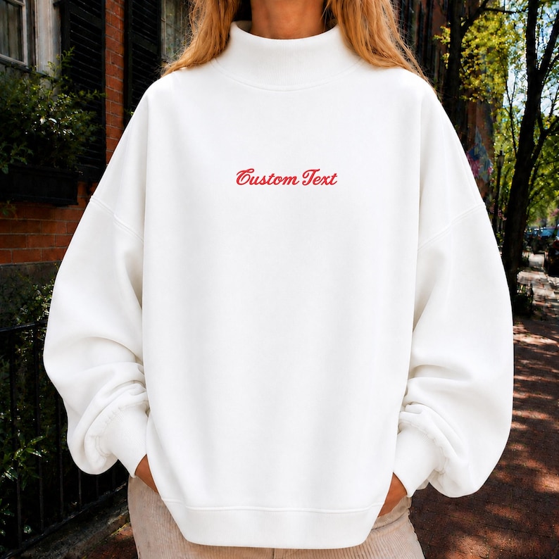 May include: An oversized white sweatshirt featuring a mock neck collar and long sleeves with ribbed cuffs. The center of the chest displays the words Custom Text embroidered in a red cursive script font, providing a clear and simple design detail.