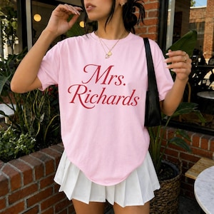 Może przedstawiać: Jasnor&oacute;żowy t-shirt z kr&oacute;tkim rękawem z napisem Mrs. Richards czerwoną czcionką szeryfową na klatce piersiowej, zestawiony z białą plisowaną minisp&oacute;dniczką i czarną torebką na ramię z cienkim paskiem.