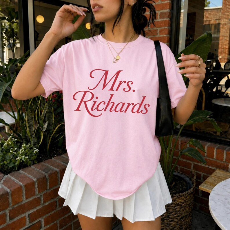Może przedstawiać: Jasnor&oacute;żowy t-shirt z kr&oacute;tkim rękawem z napisem Mrs. Richards czerwoną czcionką szeryfową na klatce piersiowej, zestawiony z białą plisowaną minisp&oacute;dniczką i czarną torebką na ramię z cienkim paskiem.