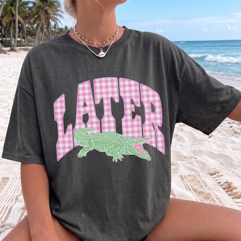 May include: Dark gray t-shirt with the word "LATER" in pink gingham print and a green alligator graphic. The shirt is a relaxed fit, perfect for casual wear.