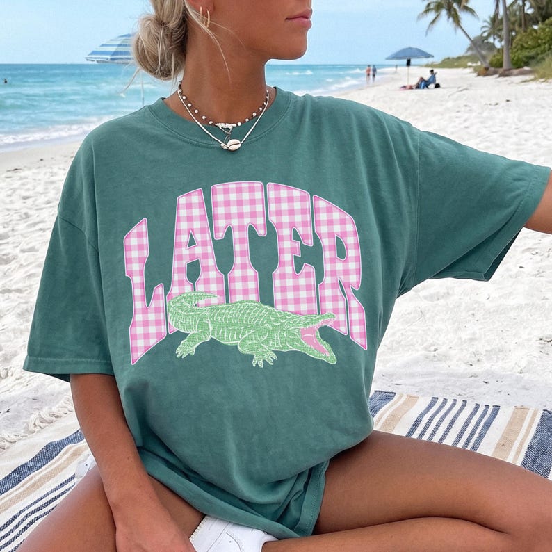May include: Green t-shirt with the word "LATER" in pink and white checkered letters and a green alligator graphic. The shirt is a relaxed fit and is being worn on a beach.
