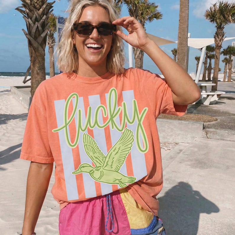 May include: An orange t-shirt with the word "lucky" in green script and a green duck graphic. The shirt has light blue vertical stripes. The person in the image is wearing sunglasses and colorful shorts.