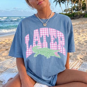 May include: A blue t-shirt with the word "LATER" in pink and white checkered letters. Below the text is a green alligator with an open mouth. The shirt is worn on a beach.