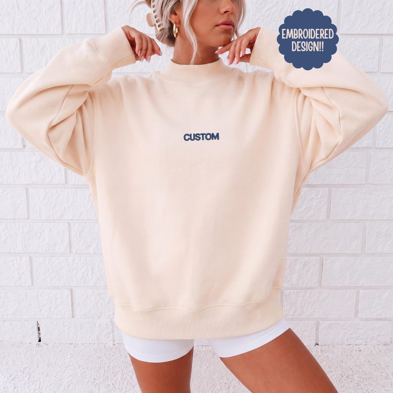 May include: A cream-colored long-sleeve sweatshirt featuring a mock neck and ribbed cuffs. The word CUSTOM is embroidered in dark blue thread on the center chest. A dark blue circular badge in the upper right corner reads EMBROIDERED DESIGN!!