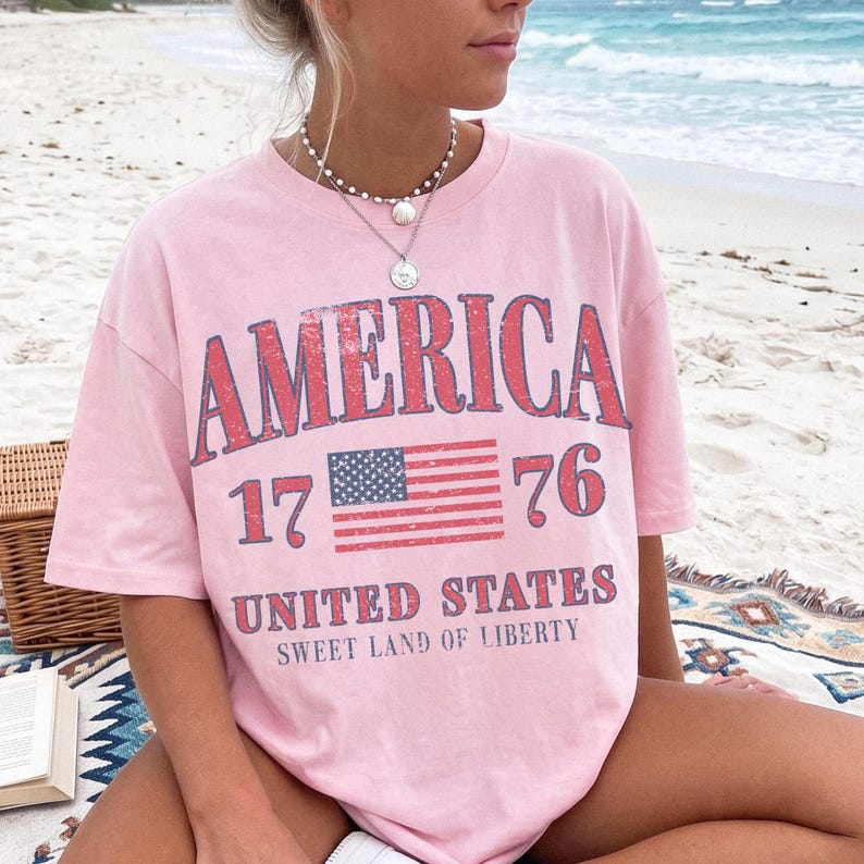 May include: A pale pink t-shirt featuring the word "AMERICA" in red, the numbers "1776", and an American flag design. The shirt also displays the text "UNITED STATES" and "SWEET LAND OF LIBERTY".