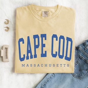 May include: A light yellow t-shirt with the words "CAPE COD" in blue block letters above "MASSACHUSETTS". The shirt is folded and placed on a white surface next to a pair of gold hoop earrings, a hair clip, and a pair of blue jeans.